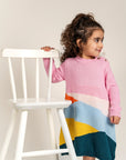 Toddler wearing Serenity Skies Cotton Knitted Sweater Dress featuring pink sleeves and colorful geometric patterns in yellow, blue, orange, and green. Soft and cozy, perfect for fall/winter casual wear.