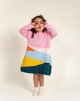 Toddler wearing Serenity Skies Cotton Knitted Sweater Dress with pink sleeves and vibrant geometric patterns in yellow, blue, orange, and green. Soft, cozy cotton perfect for playful fall/winter outfits.