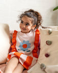 Toddler sitting on a couch wearing the Petal Playtime Cotton Knitted Dress with pink sleeves, orange ruffles, and a blue flower design. Soft, cozy, perfect for casual fall/winter outfits.