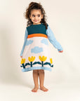 Toddler girl wearing Cloudy Daydream Cotton Knitted Sweater Dress with blue sleeves, cloud and yellow tulip patterns, and orange stripe. Cozy, long-sleeved cotton perfect for casual fall/winter outfits.