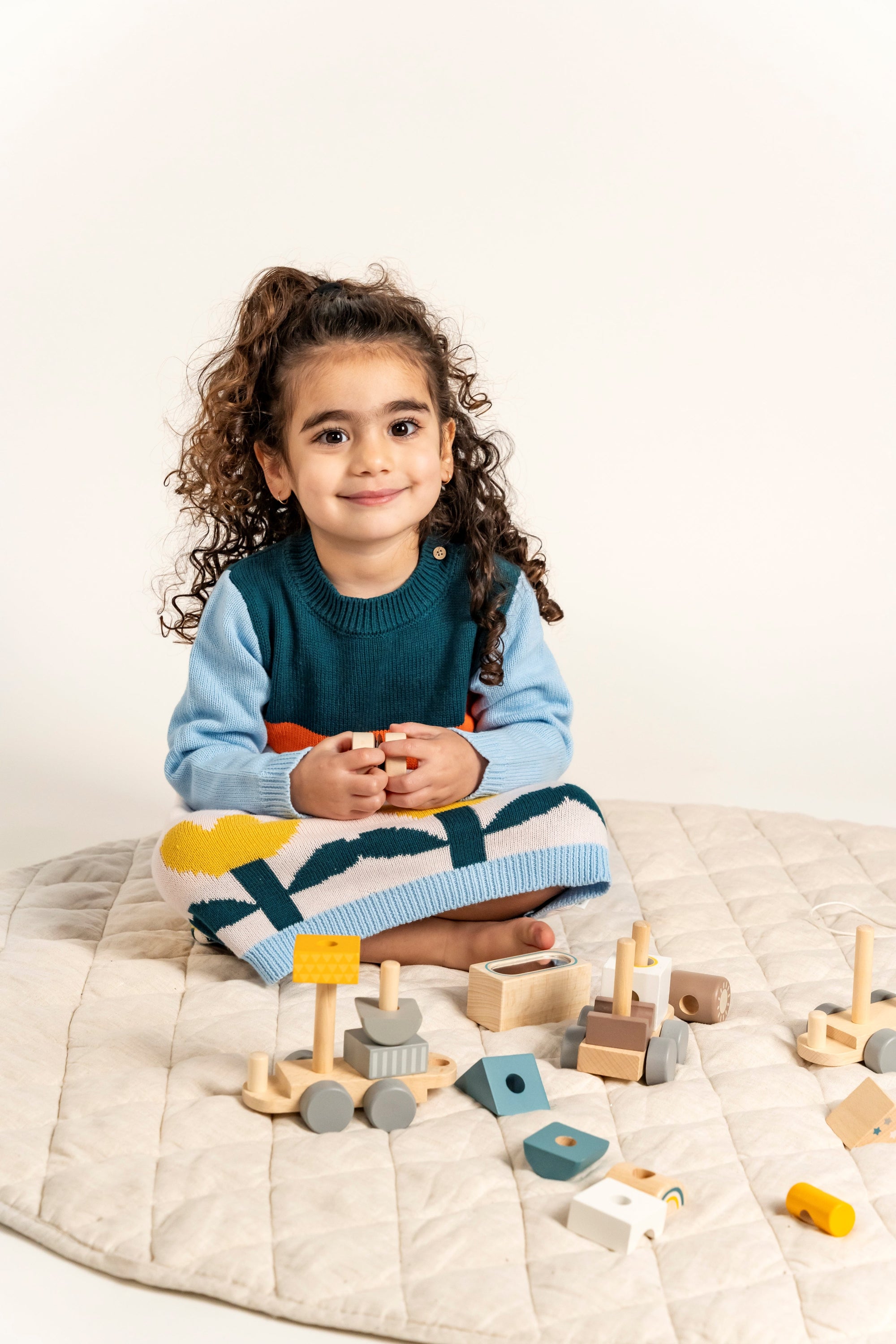 Toddler sitting and playing while wearing the Cloudy Daydream Cotton Knitted Sweater Dress, featuring blue sleeves, cloud patterns, and yellow tulips. Soft and cozy cotton, perfect for cooler seasons.