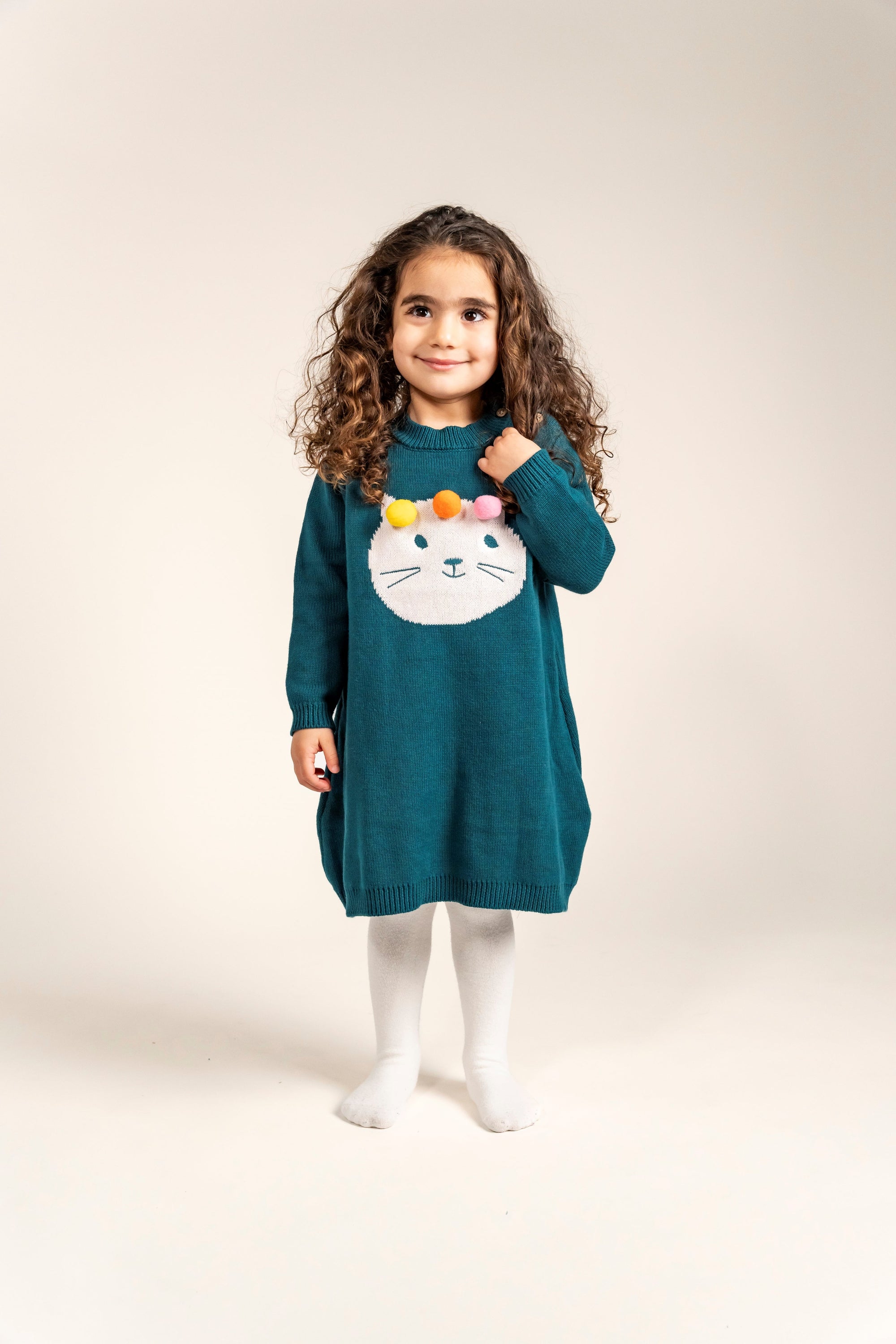 Toddler wearing Pompom Wonderland Cotton Knitted Sweater Dress in dark green with a white cat design and colorful pom-poms. Long-sleeved, cozy, soft cotton ideal for casual fall and winter outfits.