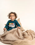 Toddler sitting wrapped in a cozy blanket wearing Pompom Wonderland Cotton Knitted Sweater Dress in dark green with a white cat design and colorful pom-poms. Soft, long-sleeved, perfect for fall/winter wear.