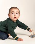 Adorable baby wearing a forest green Waffle Cotton Knit Jumper with yellow and white stripe details on the pocket and cuffs, playing with a wooden toy. Ideal cozy sweater for your little one’s everyday adventures.