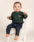 Cute toddler wearing a forest green Waffle Cotton Knit Jumper with yellow and white stripe details on the pocket and cuffs, seated on a white chair. Cozy and stylish, perfect for everyday wear.