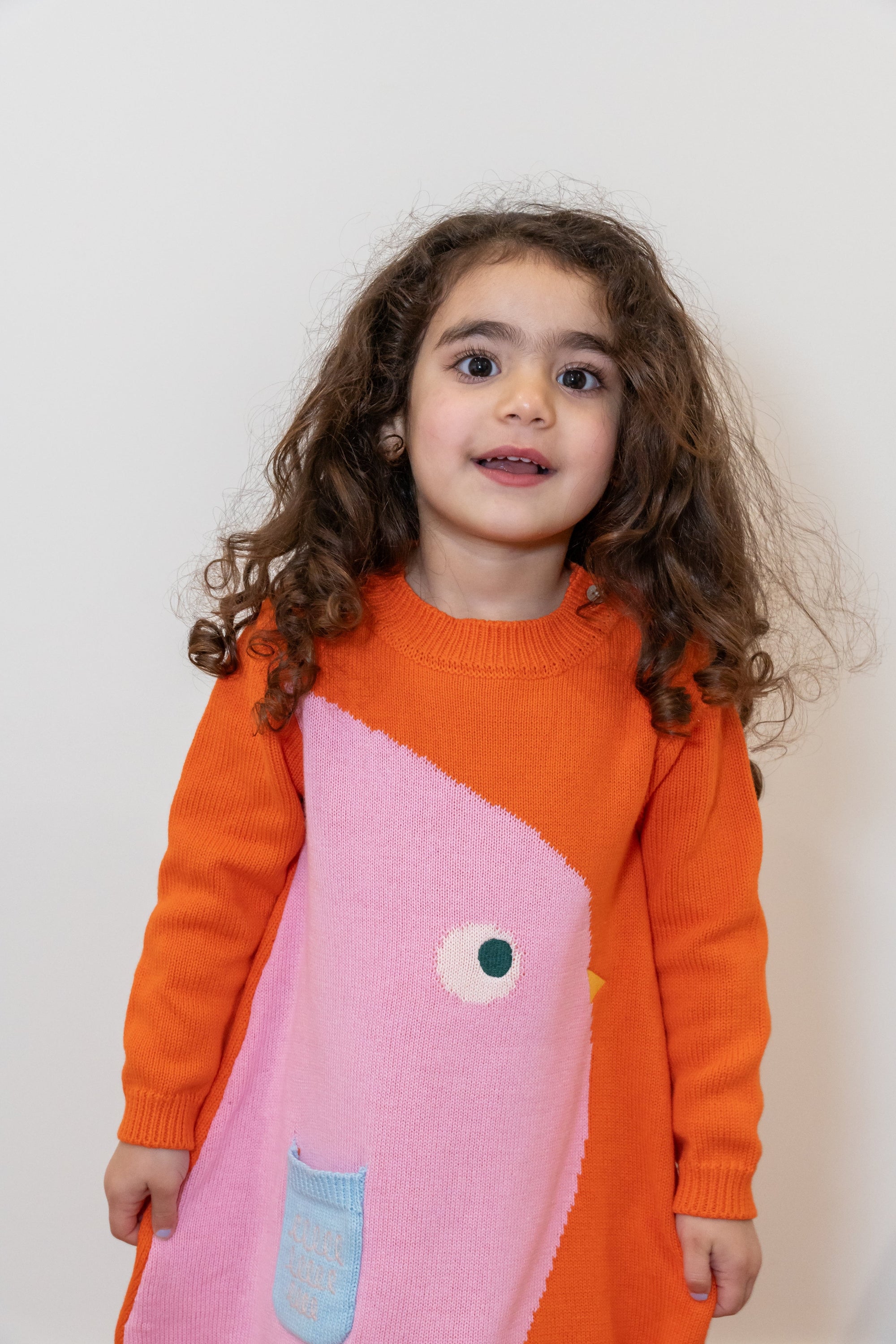 Toddler wearing a Chirpy Bird Cotton Knitted Sweater Dress in orange with a pink bird design and front pocket. Soft, long-sleeved, cozy cotton fabric perfect for fall and winter. Ideal for casual outings.