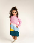 Toddler wearing Serenity Skies Cotton Knitted Sweater Dress with pink sleeves and colorful geometric patterns in blue, yellow, orange, and green. Soft, cozy, perfect for casual fall and winter wear.